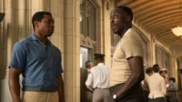 Dois homens negros conversam intensamente num salão com arquitetura clássica. Um veste camisa azul, outro bege. O clima é sério.