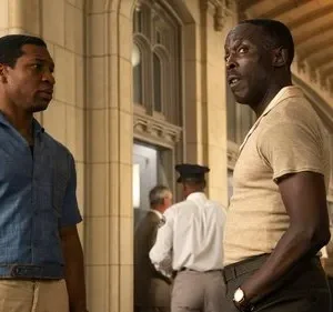 Dois homens negros conversam intensamente num salão com arquitetura clássica. Um veste camisa azul, outro bege. O clima é sério.