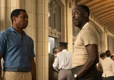 Dois homens negros conversam intensamente num salão com arquitetura clássica. Um veste camisa azul, outro bege. O clima é sério.