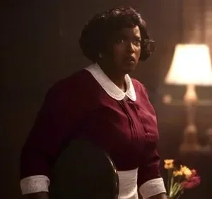 Mulher negra de cabelo curto e escuro, em uniforme bordô e branco, olha apreensiva. Fundo escuro com abajur iluminado, criando clima tenso.