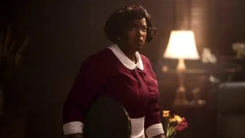 Mulher negra de cabelo curto e escuro,em uniforme bordô e branco,olha apreensiva. Fundo escuro com abajur iluminado,criando clima tenso.
