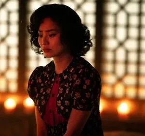 Mulher de cabelo escuro, com expressão pensativa, em ambiente com luz de velas e fundo de padrão geométrico. Clima introspectivo.