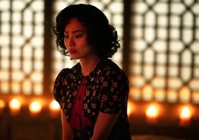 Mulher de cabelo escuro, com expressão pensativa, em ambiente com luz de velas e fundo de padrão geométrico. Clima introspectivo.