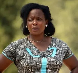 Mulher negra com cabelo escuro, vestido estampado em cinza e azul, expressa seriedade. Fundo natural e desfocado.