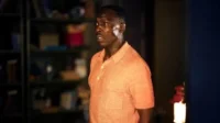 Homem negro de camisa laranja, com olhar surpreso e boca aberta. Está em um ambiente interno mal iluminado.