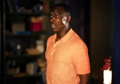 Homem negro de camisa laranja, com olhar surpreso e boca aberta. Está em um ambiente interno mal iluminado.