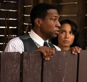 Um homem e uma mulher olham apreensivos por cima de uma cerca de madeira. Ele usa colete, ela tem cabelo ondulado. Cenário rústico.