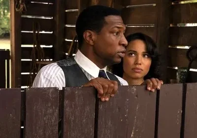 Um homem e uma mulher olham apreensivos por cima de uma cerca de madeira. Ele usa colete, ela tem cabelo ondulado. Cenário rústico.