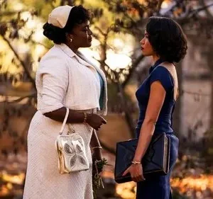 Duas mulheres com figurinos de época se encaram num parque com luz quente de outono. Uma veste creme, a outra azul-marinho. Atmosfera séria.