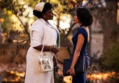 Duas mulheres com figurinos de época se encaram num parque com luz quente de outono. Uma veste creme, a outra azul-marinho. Atmosfera séria.