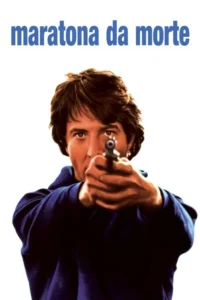 Homem com cabelo castanho e camisa azul escura aponta uma arma para a câmera,com olhar intenso,em fundo branco.