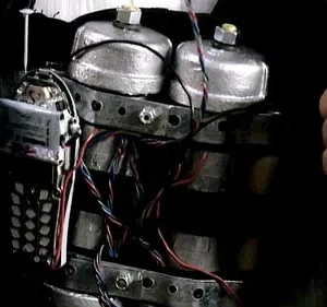 Duas mãos dão polegares para cima ao lado de um dispositivo explosivo improvisado com um celular e muitos fios, no peito de alguém.