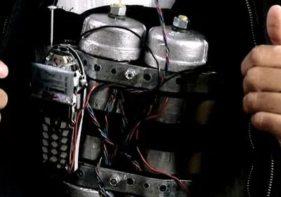 Duas mãos dão polegares para cima ao lado de um dispositivo explosivo improvisado com um celular e muitos fios, no peito de alguém.