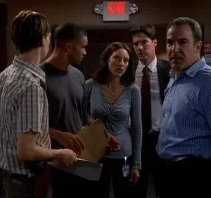 Seis pessoas em um escritório. Um homem de camisa azul listrada no primeiro plano olha sério para frente, enquanto o grupo parece tenso.