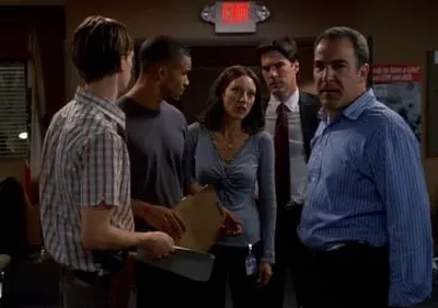 Seis pessoas em um escritório. Um homem de camisa azul listrada no primeiro plano olha sério para frente, enquanto o grupo parece tenso.