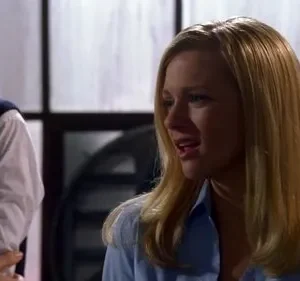 Uma mulher loira de camisa azul claro em primeiro plano parece estar falando. Um homem de colete azul escuro está atrás. Cena interna profissional.