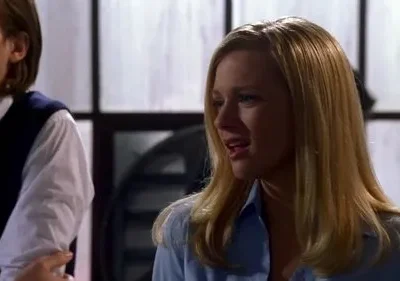 Uma mulher loira de camisa azul claro em primeiro plano parece estar falando. Um homem de colete azul escuro está atrás. Cena interna profissional.