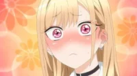 Garota anime loira, olhos rosa arregalados e rosto corado de vergonha, contra fundo floral. Parece surpresa.