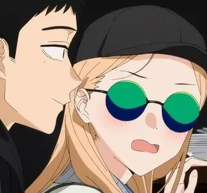 Um homem sussurra para uma mulher loira de boné e óculos de sol, que está corada e com expressão surpresa. Taças douradas ao fundo.