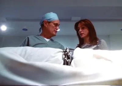 Um cirurgião e uma mulher séria olham para baixo sobre um lençol branco em sala médica. Clima sério.