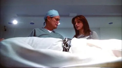 Um cirurgião e uma mulher séria olham para baixo sobre um lençol branco em sala médica. Clima sério.