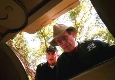 Dois homens, um de chapéu e outro de boné com NCIS, olham para baixo em uma abertura ao ar livre, com árvores ao fundo. Clima investigativo.