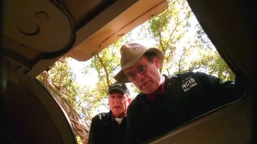 Dois homens,um de chapéu e outro de boné com NCIS,olham para baixo em uma abertura ao ar livre,com árvores ao fundo. Clima investigativo.