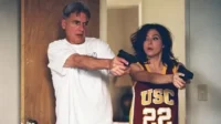 Homem grisalho e mulher em regata USC,ambos com pistolas e expressões de surpresa/susto,ela segurando duas. Cenário interno.