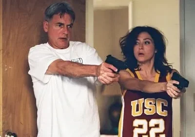 Homem grisalho e mulher em regata USC, ambos com pistolas e expressões de surpresa/susto, ela segurando duas. Cenário interno.