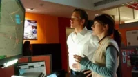 Duas pessoas focadas num centro de operações. Um homem e uma mulher observam um grande monitor com um mapa digital. Clima de tensão.