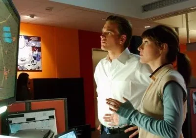 Duas pessoas focadas num centro de operações. Um homem e uma mulher observam um grande monitor com um mapa digital. Clima de tensão.