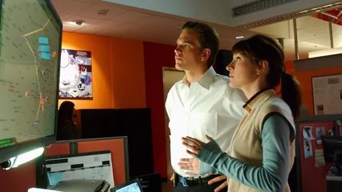 Duas pessoas focadas num centro de operações. Um homem e uma mulher observam um grande monitor com um mapa digital. Clima de tensão.