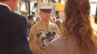 Um militar em uniforme bege e boné branco fala em vários microfones. Repórteres e uma multidão assistem em uma rua ensolarada.