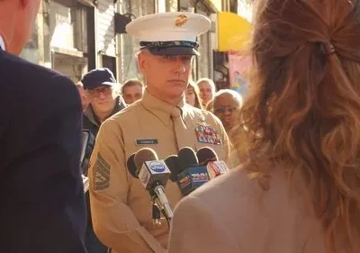 Um militar em uniforme bege e boné branco fala em vários microfones. Repórteres e uma multidão assistem em uma rua ensolarada.