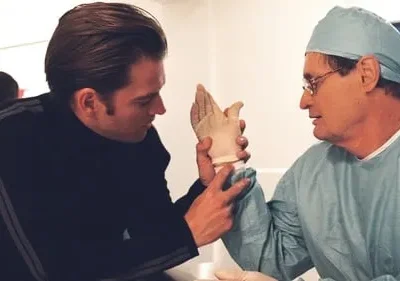 Jovem em casaco preto mostra uma luva protética a um cirurgião de óculos e touca azul. Cena séria em ambiente clínico.