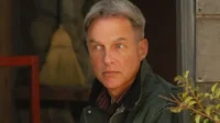 NCIS:Investigação Naval – T01E15:Enigma 8 Um homem de meia-idade de cabelo grisalho e jaqueta verde olha intensamente para o lado,num cenário rústico com folhagem.