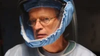 NCIS:Investigação Naval – T01E16:Nêmeses 7 Um homem de óculos,vestindo um capacete azul com visor e avental médico,olha fixamente em um ambiente escuro e sério.