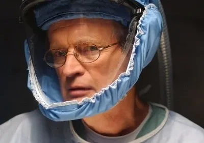 Um homem de óculos, vestindo um capacete azul com visor e avental médico, olha fixamente em um ambiente escuro e sério.