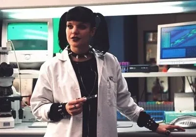 Mulher de cabelo preto com visual alternativo, em jaleco de laboratório, cercada por equipamentos científicos. Segura um objeto.