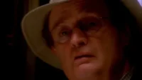 NCIS:Investigação Naval – T01E19:O Homem Morto Falou 4 Close-up de um homem de óculos e chapéu claro,rosto iluminado por uma luz laranja na escuridão,expressão preocupada.