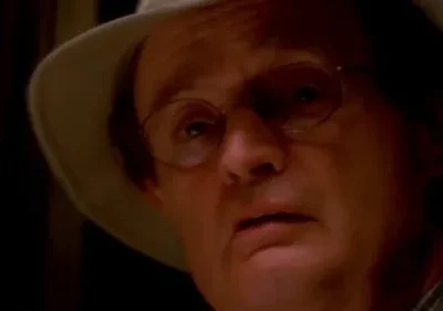 Close-up de um homem de óculos e chapéu claro, rosto iluminado por uma luz laranja na escuridão, expressão preocupada.