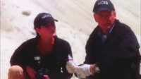 NCIS:Investigação Naval – T01E22:O Elo Fraco 2 Dois agentes de boné escuro inspecionam evidências em um local árido. O homem de luvas segura um saco,com foco sério.