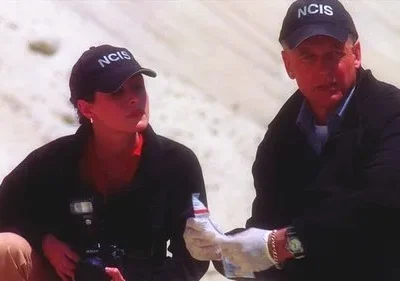 Dois agentes de boné escuro inspecionam evidências em um local árido. O homem de luvas segura um saco, com foco sério.