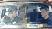 Dois rapazes num carro antigo. Um, de cabelo cacheado, olha sério; o outro, com sardas, sorri. Ambiente simples e íntimo.