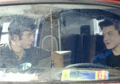 Dois rapazes num carro antigo. Um, de cabelo cacheado, olha sério; o outro, com sardas, sorri. Ambiente simples e íntimo.
