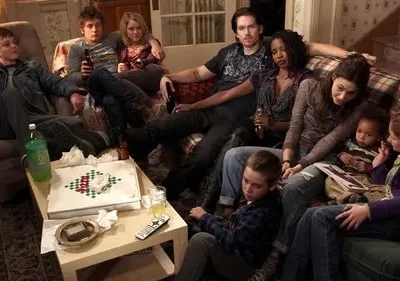 Um grupo diverso de adultos e crianças relaxa em uma sala de estar desordenada com pizza e bebidas. Clima familiar, caótico e acolhedor.