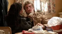 Mulher com dreads cuida de homem com rosto machucado e nariz enfaixado,usando luvas. Suprimentos médicos em mesa doméstica. Cena de cuidado.