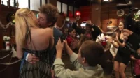 Casal se beija apaixonadamente em festa,enquanto outros dançam e celebram. Um garoto filma a cena. Ambiente festivo e alegre.