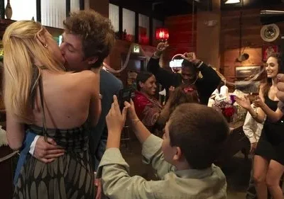Casal se beija apaixonadamente em festa, enquanto outros dançam e celebram. Um garoto filma a cena. Ambiente festivo e alegre.