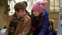 Jovem e garota em roupas de inverno. Ele de jaqueta marrom e cachecol,ela com gorro rosa e casaco azul. Ambiente interno,clima pensativo.